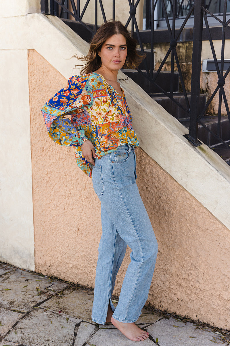 Woman in a colorful floral blouse and blue jeans standing against a textured wall.