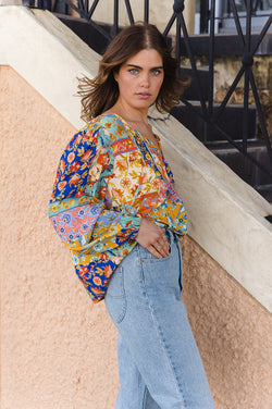 Woman wearing a colorful patterned blouse and blue jeans standing against a textured wall.