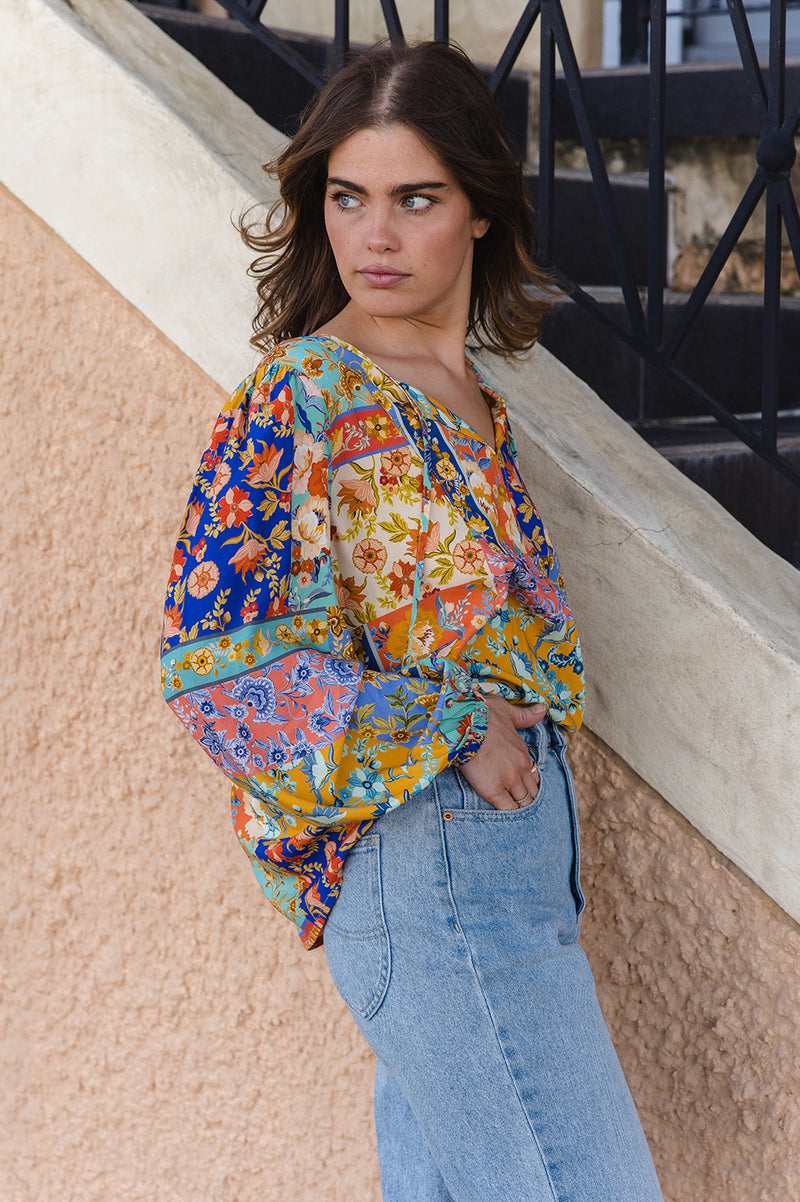 Woman wearing a colorful patterned blouse and blue jeans standing against a textured wall.