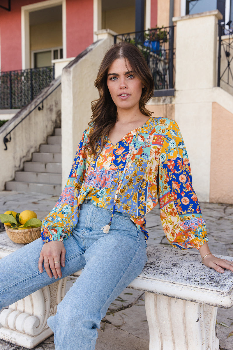 Woman sitting on a stone bench wearing a colorful floral blouse and blue jeans, with a building and steps in the background.
