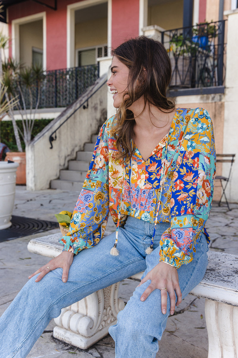 Woman sitting on a bench wearing a colorful floral blouse and blue jeans.