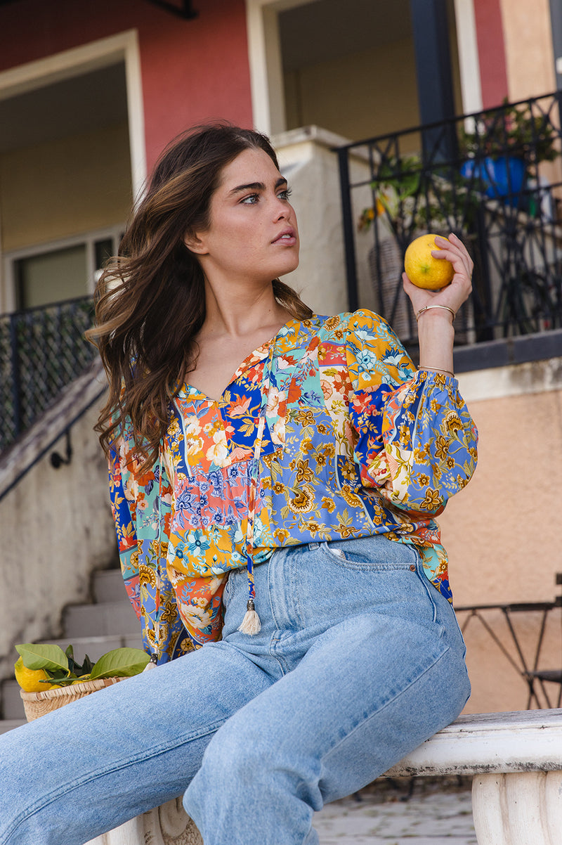 Woman in a colorful floral blouse and jeans holding an orange, sitting on steps.