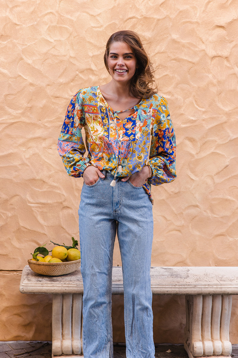 Woman wearing a colorful floral blouse and blue jeans standing against a textured beige wall.
