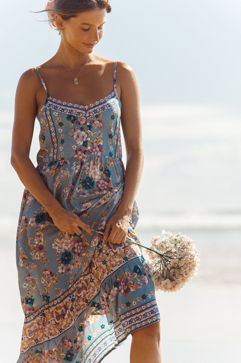 Brunette woman, standing on the beach and wearing a floral printed midi length dress. The dress has a frilled hemline with border detailing and there is placement print detailing throughout the dress. The girl is standing on the beach looking down at her Hands. she is holding a hydrangea 