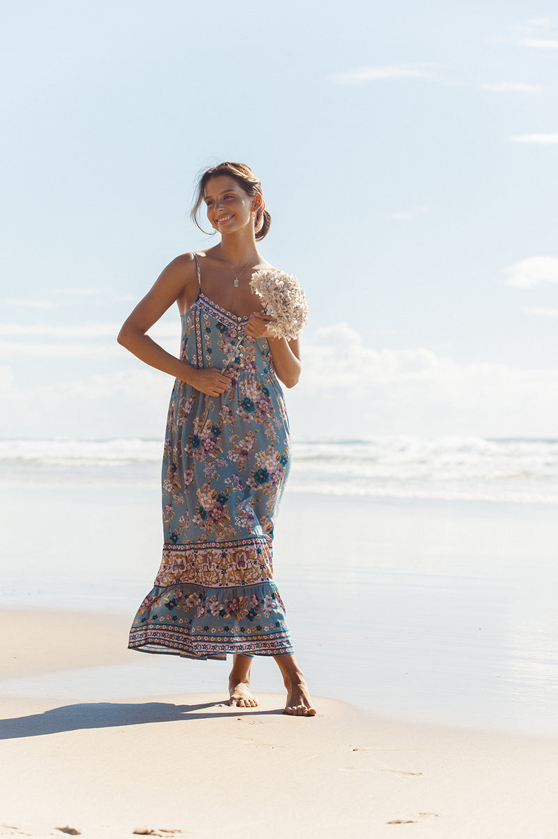 A brunette woman is standing on the beach with her legs crossed and is holding her arms up near her chest while holding a long stemmed white hydrangea. She has her legs crossed and is wearing a midi length dress in a blue floral print with placement print detailing