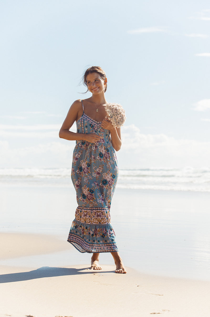 A brunette woman is standing on the beach with her legs crossed and is holding her arms up near her chest while holding a long stemmed white hydrangea. She has her legs crossed and is wearing a midi length dress in a blue floral print with placement print detailing