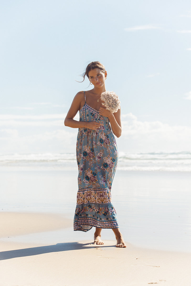 A brunette woman is standing on the beach with her legs crossed and is holding her arms up near her chest while holding a long stemmed white hydrangea. She has her legs crossed and is wearing a midi length dress in a blue floral print with placement print detailing