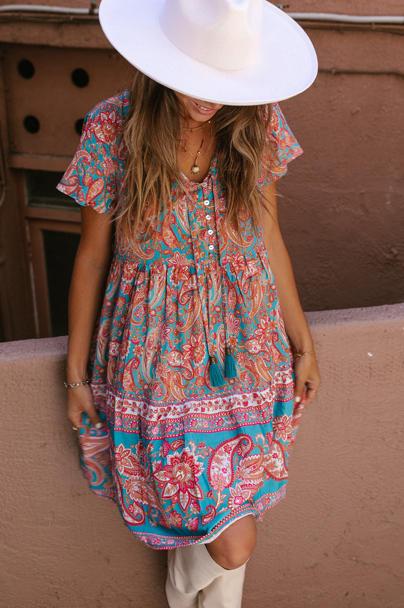 A girl with medium length brown hair and tanned skin, wearing a short sleeve mini dress. The dress is a paisley print in teal and multi colours