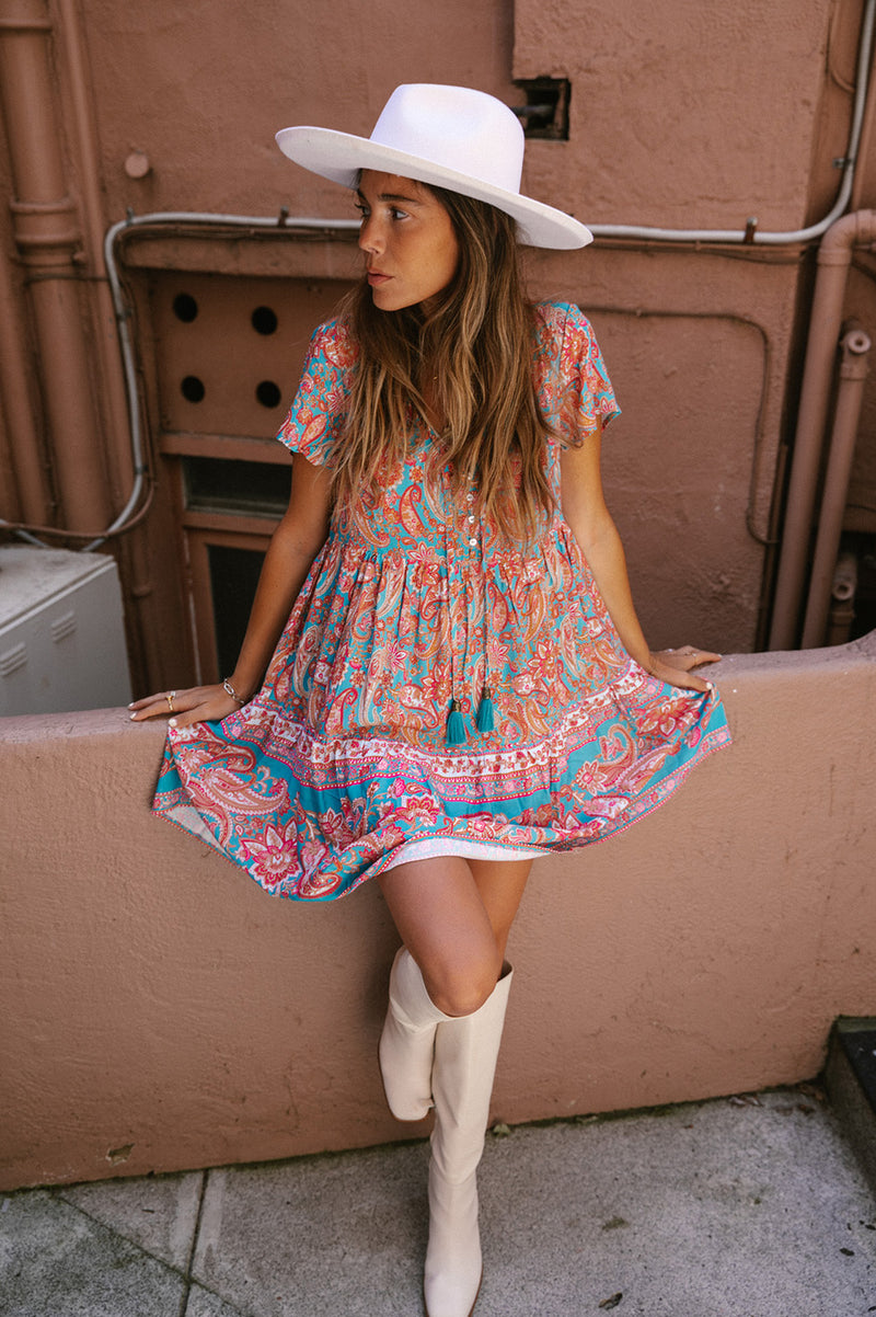 A girl with medium length brown hair and tanned skin, wearing a short sleeve mini dress. The dress is a paisley print in teal and multi colours